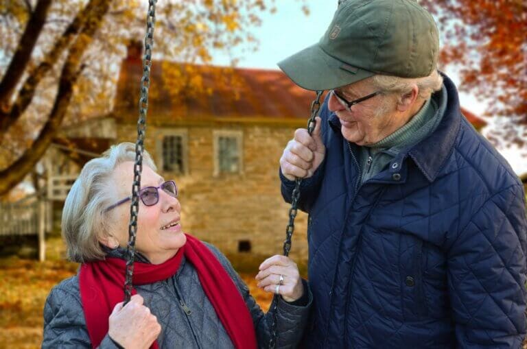 senior retired man standing next to senior retired woman sitting on swing - retirement pension providing well